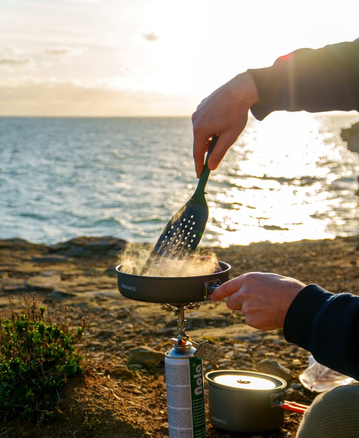 Hände kochen Essen auf einem Campingkocher am Meer.